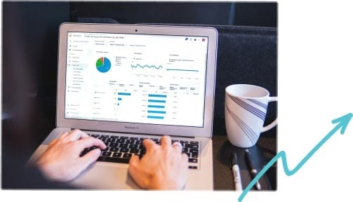 Person working on a laptop displaying analytics graphs and charts, with a coffee mug and pens beside them. An upward arrow graphic is visible in the lower right corner, symbolizing growth or progress.