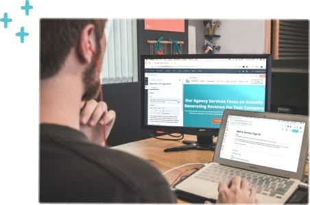 A person sits at a desk using a laptop and an external monitor. The monitor displays a website about agency services and revenue generation, while the laptop shows a text document.
