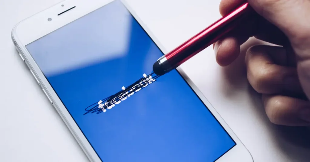 The image shows a smartphone with the Facebook logo displayed on the screen. The logo is crossed out with black ink, as if someone has scribbled over it. A hand holding a stylus is visible, with the stylus hovering over the scribbled-out logo, suggesting an action of erasing or rejecting Facebook.
