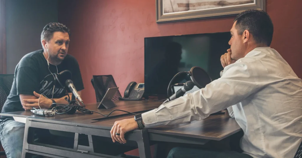Two men are sitting at a table in an office setting, engaged in a conversation. Each has a microphone in front of them, suggesting they are recording a podcast or an interview. One man is wearing casual clothing with earbuds, while the other is in a dress shirt. A computer monitor and office phone are visible in the background.