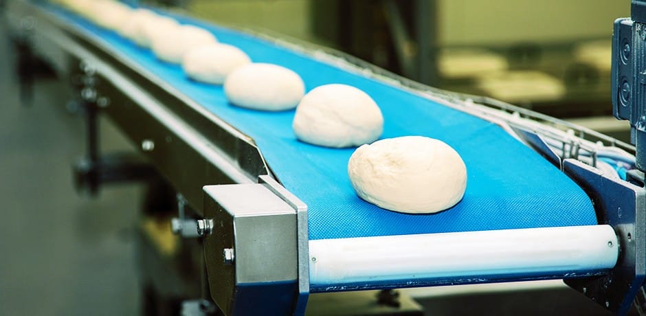 Balls of dough are evenly spaced on a blue conveyor belt in a bakery or industrial setting, ready for further processing. The background is slightly blurred, with machinery visible.