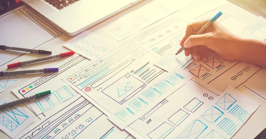A person sketches website design wireframes on multiple sheets of paper spread across a desk. There are colored pens scattered around, and a laptop is partially visible in the background. The scene is lit with natural sunlight.