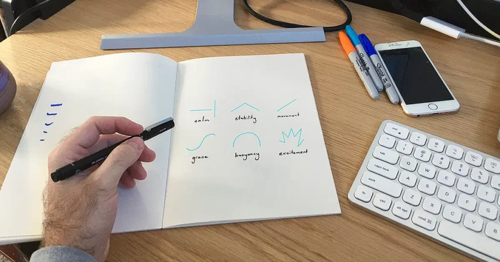 A person draws various line patterns in a notebook at a desk. Each pattern is labeled with words like "calm," "stability," "movement," "grace," "beginning," and "excitement." Nearby are a keyboard, a smartphone, and several markers, suggesting a workspace setting.