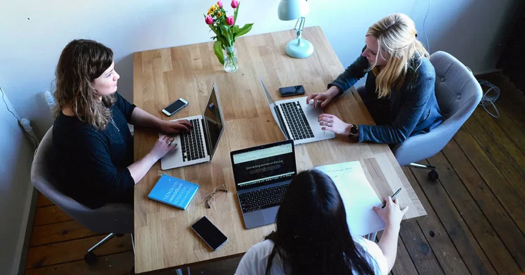 Three people sit around a wooden table using laptops. A vase of tulips and a desk lamp are on the table, along with a book and some phones.