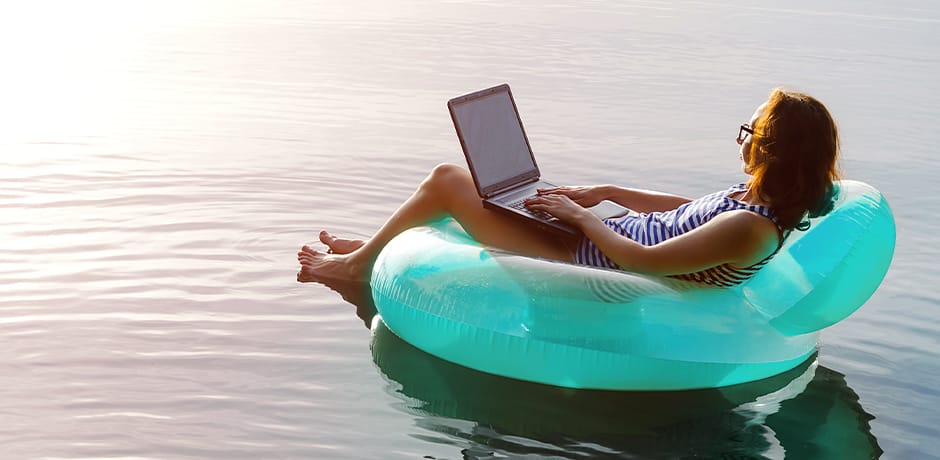 A person is relaxing on a turquoise inflatable float in a body of water while using a laptop. The sky is clear and the water is calm, creating a serene and peaceful atmosphere.