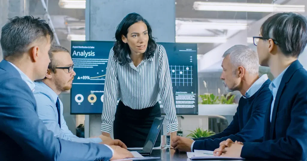A woman stands confidently at a conference table, surrounded by four seated colleagues engaged in discussion. Behind her, digital screens display charts and graphs with the word "Analysis" prominently visible. The setting is a modern office with large windows and greenery in the background.