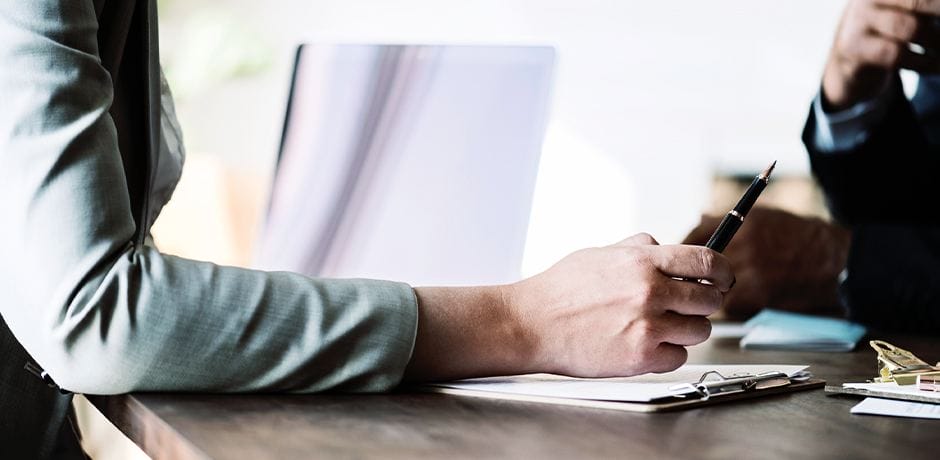 A close-up of two people at a desk, one person holding a pen and gesturing toward a clipboard with papers. A laptop is open in the background, and the scene suggests a professional meeting or discussion.