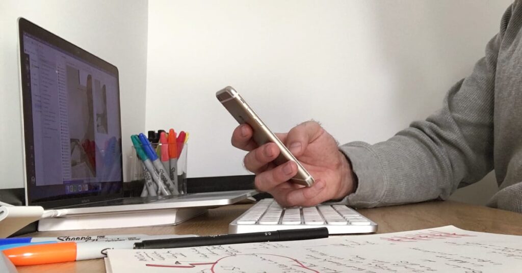 A person is sitting at a desk, holding a smartphone in one hand. On the desk are an open laptop displaying a program with a sidebar, a wireless keyboard, a notepad with handwritten notes and a diagram, and several colored markers. The setting appears to be a work or study environment.