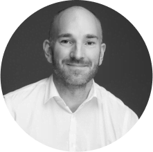 A man with a bald head and short beard, wearing a white collared shirt, smiles at the camera against a plain dark background. The image is in black and white.