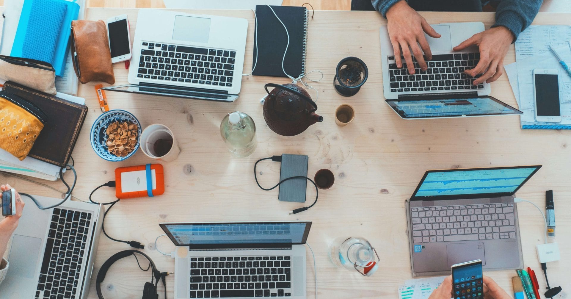 Four people working at a desk with laptops, notebooks, snacks, and gadgets.