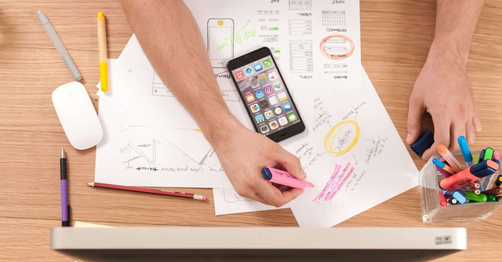Person highlighting notes on papers with a phone, pens, and computer on a wooden desk.