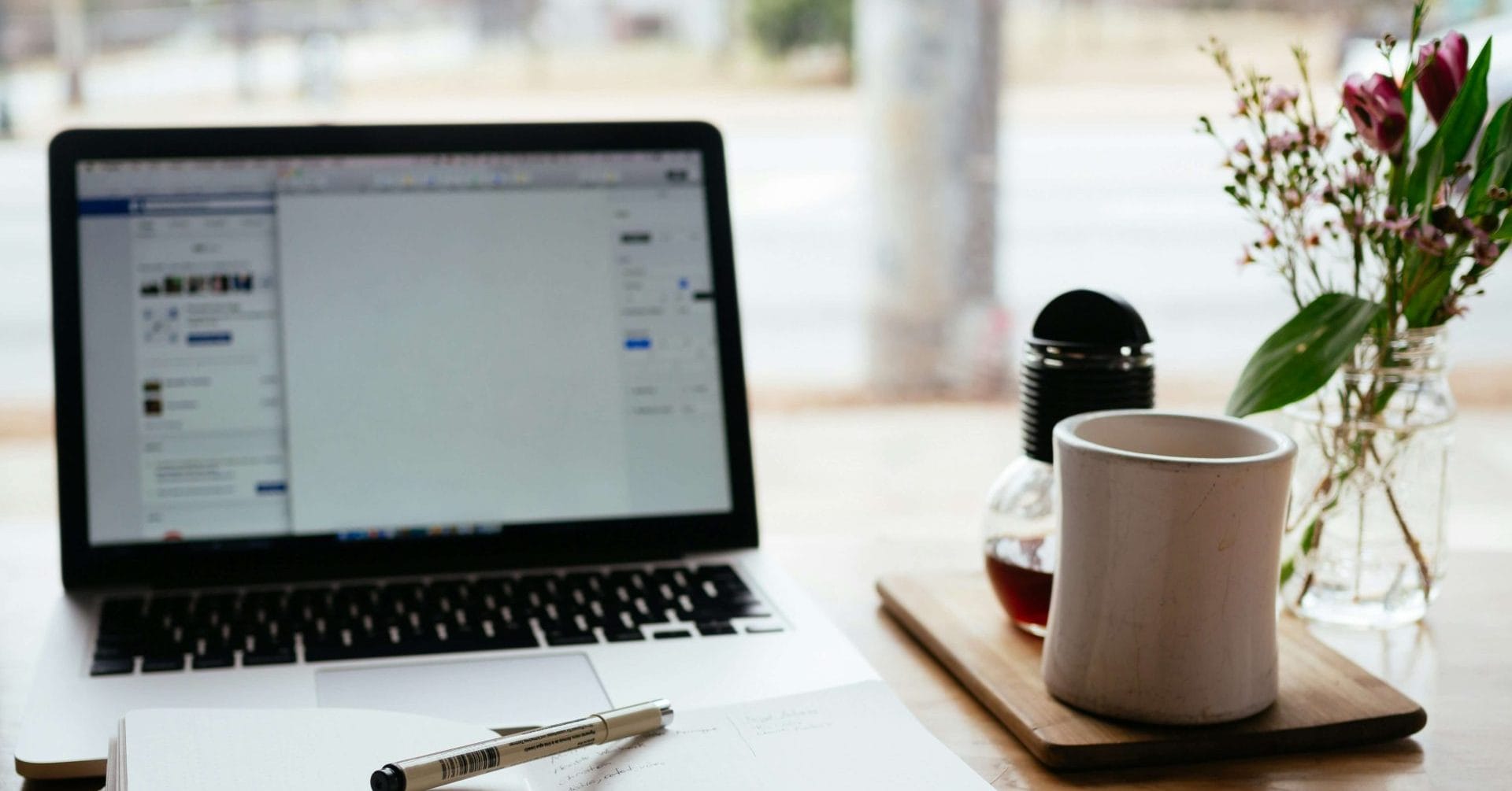Open laptop, notebook with pen, coffee mug, and flowers on a wooden table by a window.