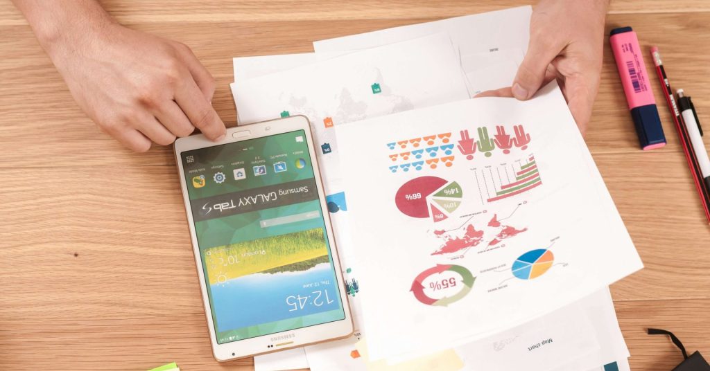 Person reviewing colorful charts on paper beside a tablet, highlighters, and sticky notes.