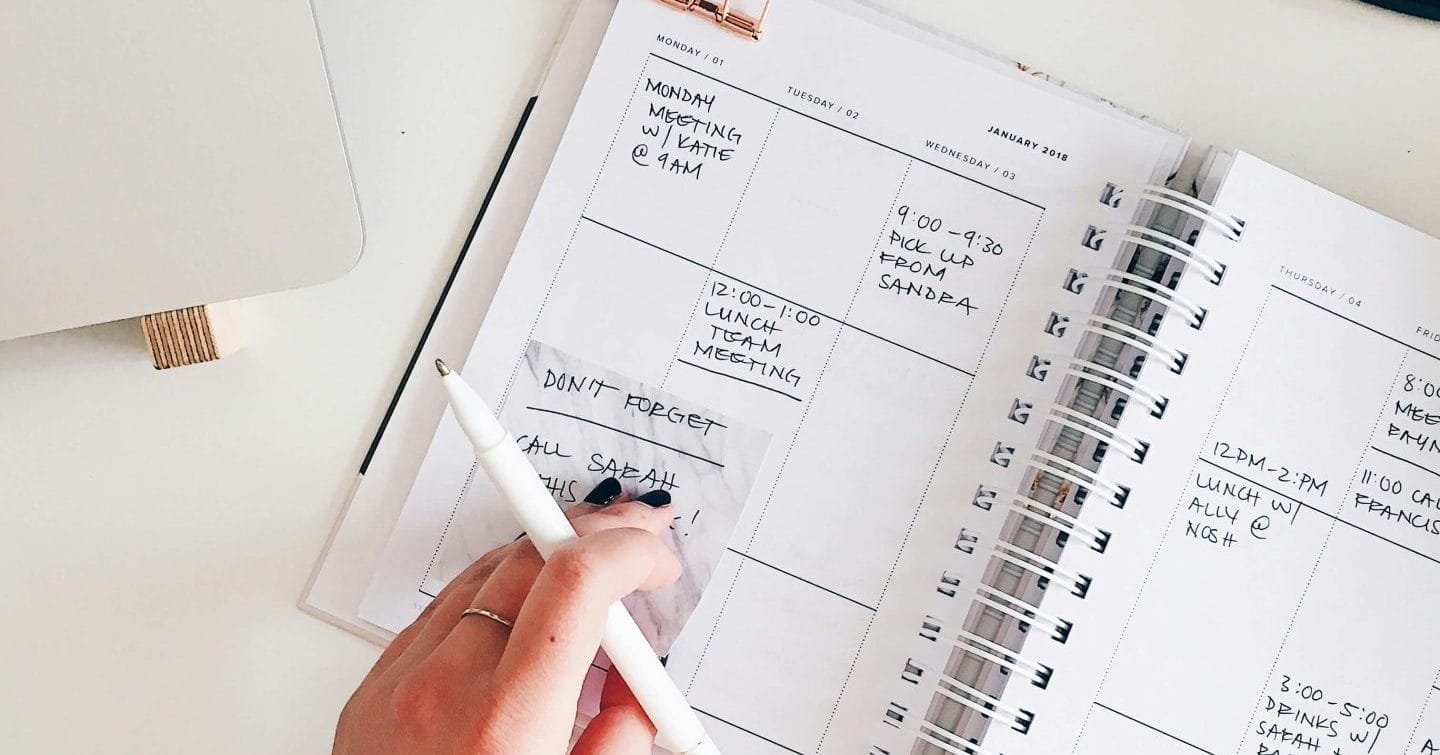 A hand writes in a planner next to a laptop, keyboard, and a cup of coffee.